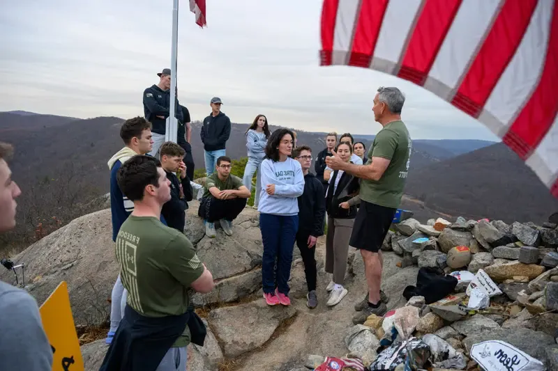 A group of people stand at the top of the Trail of the Fallen near West Point, a steep hike that ends at a large mound of stones that have been carried up and placed in memory of soldiers who have died in military conflicts. In the foreground is the American flag.